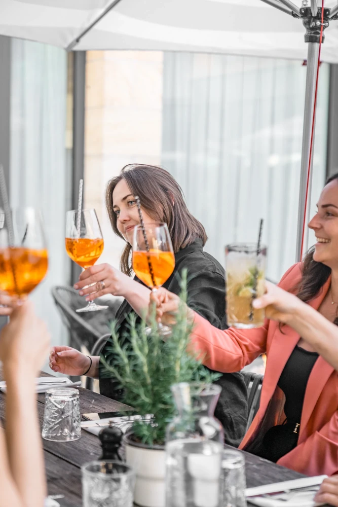 Zwei Frauen sto&szlig;en mit orangefarbenen Cocktails in einem hellen Innenraum an. Sie tragen schwarze und lachsfarbene Kleidung und sitzen an einem Tisch mit Pflanzendekoration. Die Stimmung ist fr&ouml;hlich und entspannt.