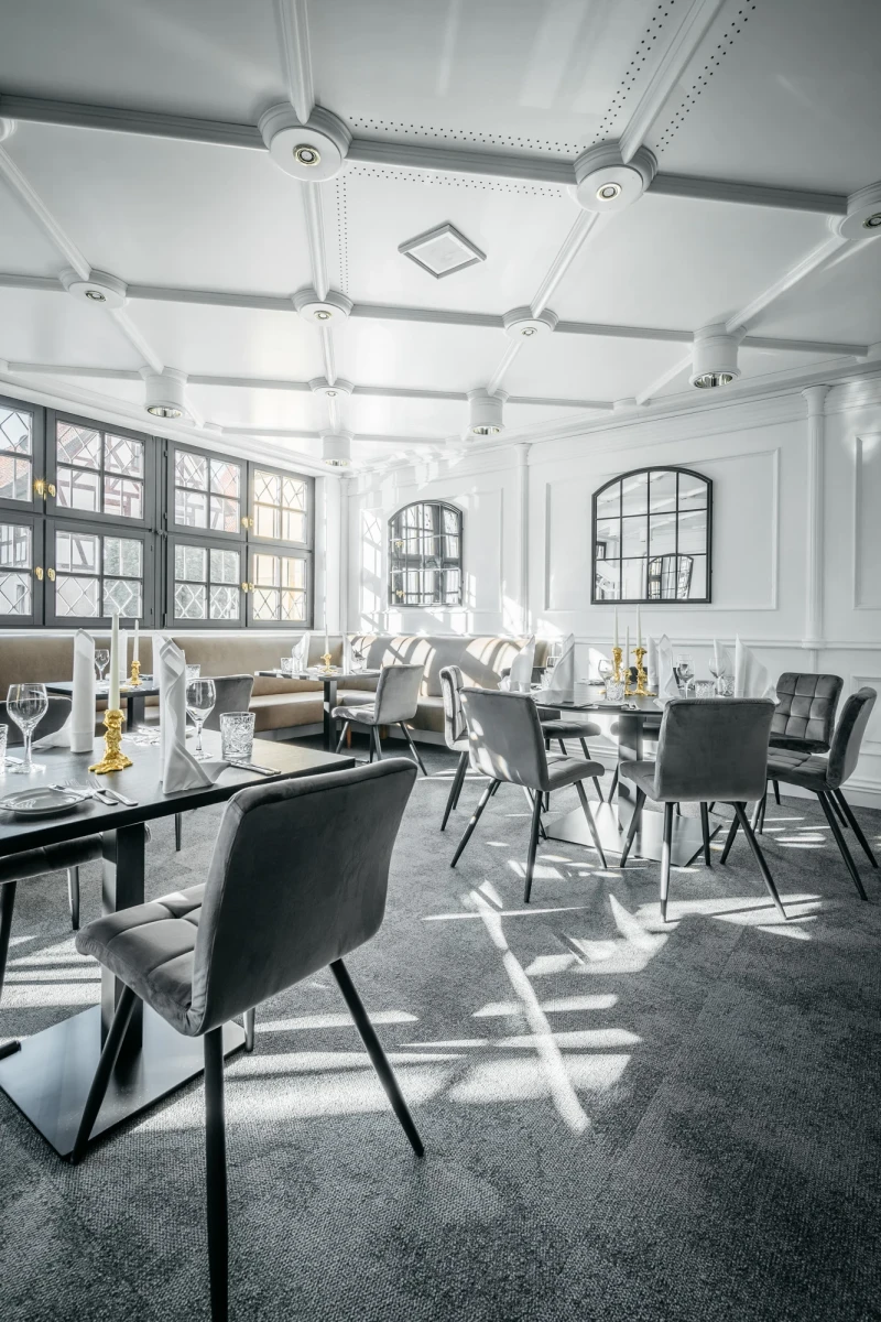 Moderner, lichtdurchfluteter Restaurantbereich mit wei&szlig;en W&auml;nden, gro&szlig;en Fenstern und grauen Designerst&uuml;hlen. Elegante Tische sind gedeckt, und der Raum strahlt eine helle, luftige Atmosph&auml;re aus.