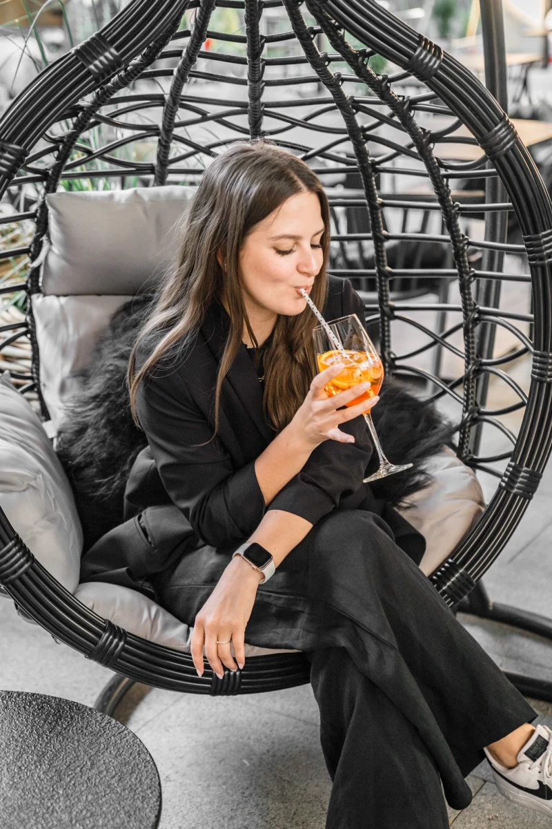 Eine Frau in schwarzer Kleidung sitzt in einem schwarzen, geschwungenen H&auml;ngesessel und h&auml;lt ein orangefarbenes Cocktailglas. Sie tr&auml;gt eine Smartwatch und scheint entspannt in einer gem&uuml;tlichen Loungeumgebung zu sein.