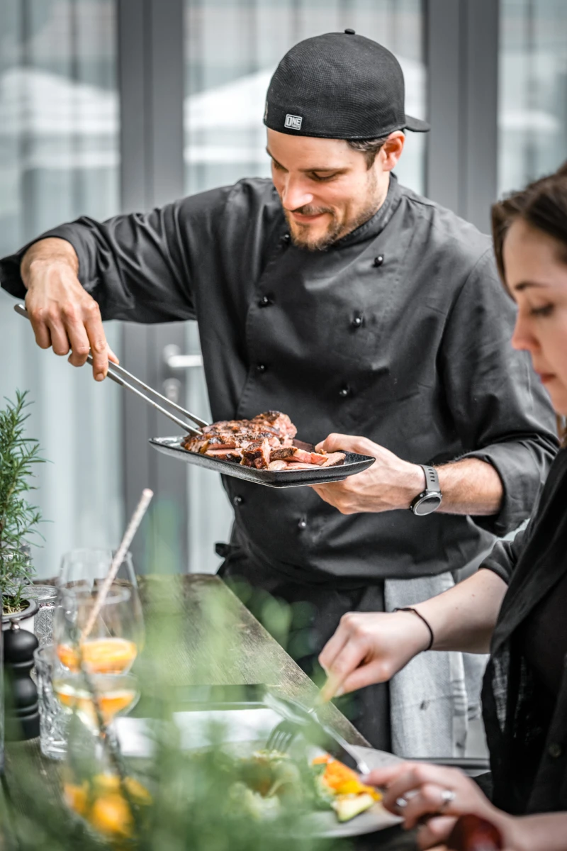 Koch in schwarzer Uniform und M&uuml;tze serviert gebratenes Fleisch von einem Teller, w&auml;hrend eine Kollegin im Hintergrund mit einer Zitrone hantiert. Professionelle K&uuml;chensituation mit appetitlich zubereiteten Speisen.