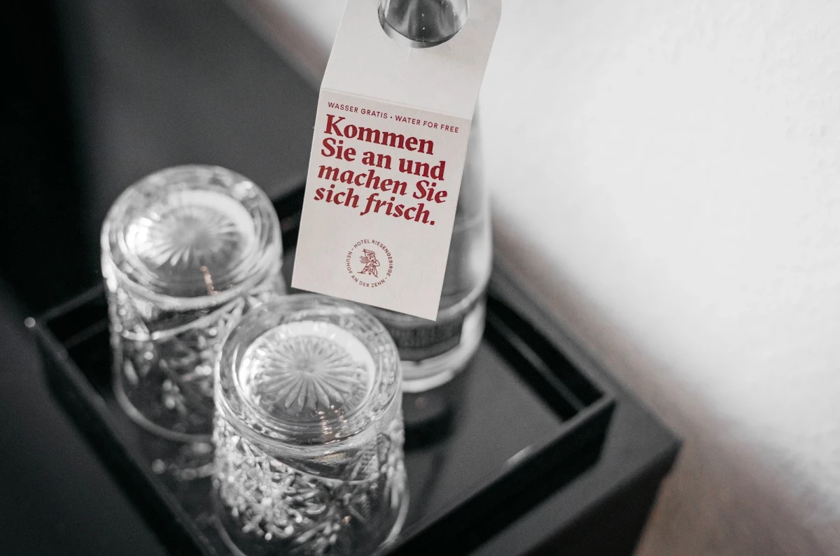 2 umgedrehte Gl&auml;ser und eine Wasserflasche stehen auf einem dunklen Tablett. An der Wasserflasche h&auml;ngt ein Papier-Anh&auml;nger mit der Aufschrift "Wasser gratis. Kommen Sie an und machen Sie sich frisch."