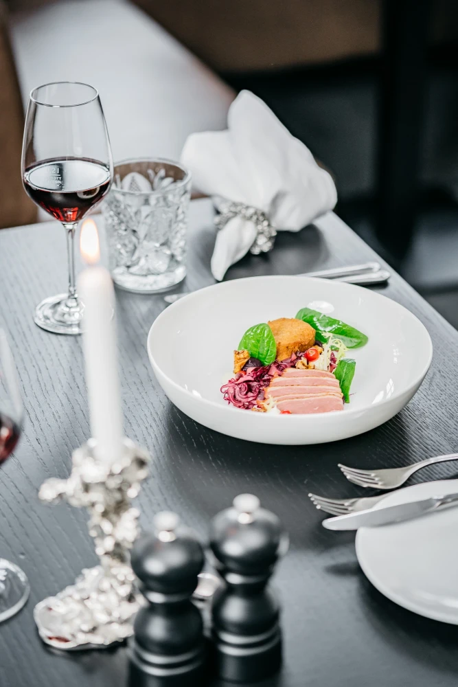 Elegantes Restaurantgedeck mit einem wei&szlig;en Teller, auf dem ein k&uuml;nstlerisch angerichtetes Gericht mit Rosa-T&ouml;nen, gr&uuml;nem Blattwerk und einer orangefarbenen Komponente serviert wird. Ein Glas Rotwein und eine brennende Kerze vervollst&auml;ndigen die stilvolle Tischdekoration.
