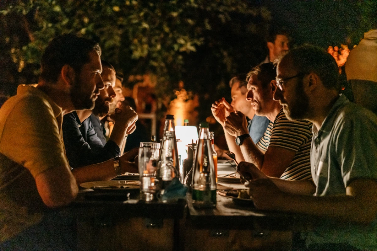 Abendliche Tischszene mit Personen, die an einem Tisch sitzen und miteinander in ged&auml;mpftem Licht kommunizieren, mit Getr&auml;nken und einer intim-geselligen Atmosph&auml;re.