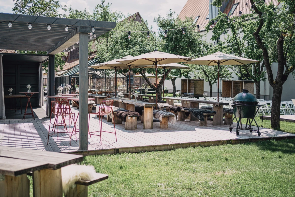 Sonnige Gartenterrasse eines Hotels mit Holztischen, wei&szlig;en Sonnenschirmen und einer &uuml;berdachten Sitzfl&auml;che. Gr&uuml;ne Rasenfl&auml;che und B&auml;ume rahmen den gem&uuml;tlichen Au&szlig;enbereich ein, der zum Verweilen einl&auml;dt.