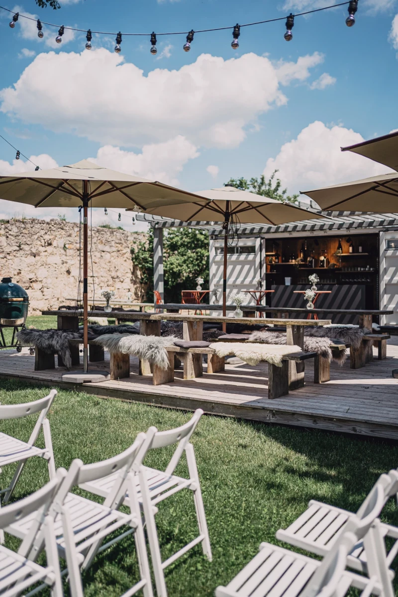 Rustikale Au&szlig;enterrasse mit Holzm&ouml;beln und wei&szlig;en Sonnenschirmen auf einem Holzdeck. H&ouml;lzerner Ausschankwagen im Hintergrund, umgeben von Rasen und Lichterketten unter blauem Himmel mit Wolken.