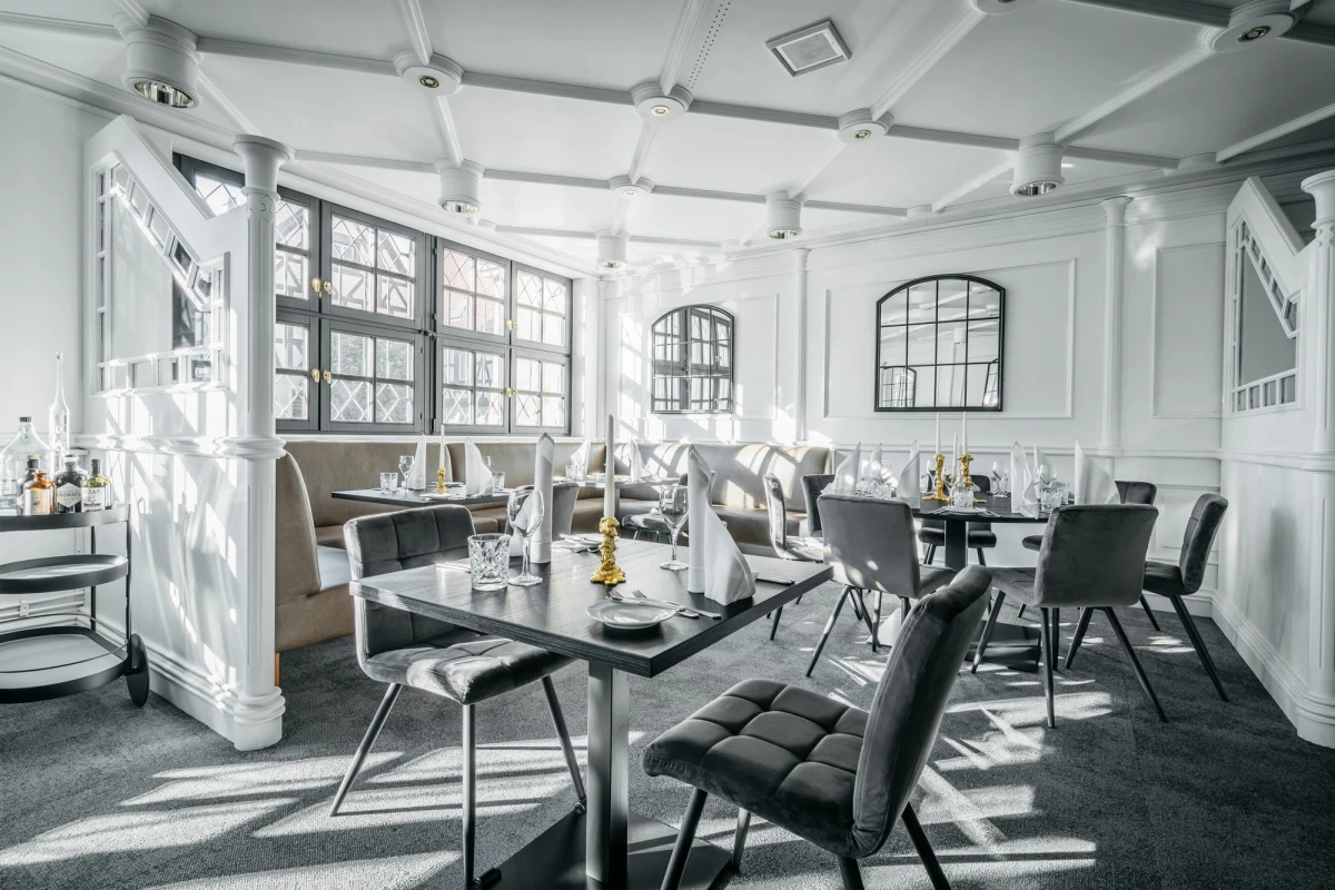 Helles, luftiges Restaurantinterieur mit modern designten St&uuml;hlen und gedeckten Tischen. Gro&szlig;e Fenster lassen viel nat&uuml;rliches Licht herein, und wei&szlig;e W&auml;nde sowie S&auml;ulen verleihen dem Raum eine elegante, offene Atmosph&auml;re.