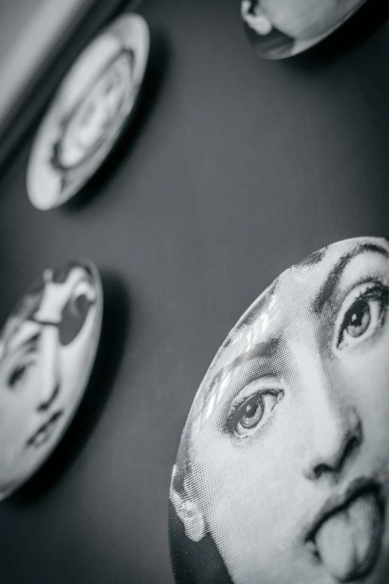 Mehrere schwarzwei&szlig;e Teller oder Keramikscheiben an der Wand, die Gesichtsteile mit detaillierten Augen und Gesichtskonturen zeigen. Die Teller sind in unterschiedlicher Unsch&auml;rfe und Perspektive angeordnet, was einen k&uuml;nstlerischen und surrealen Eindruck vermittelt.