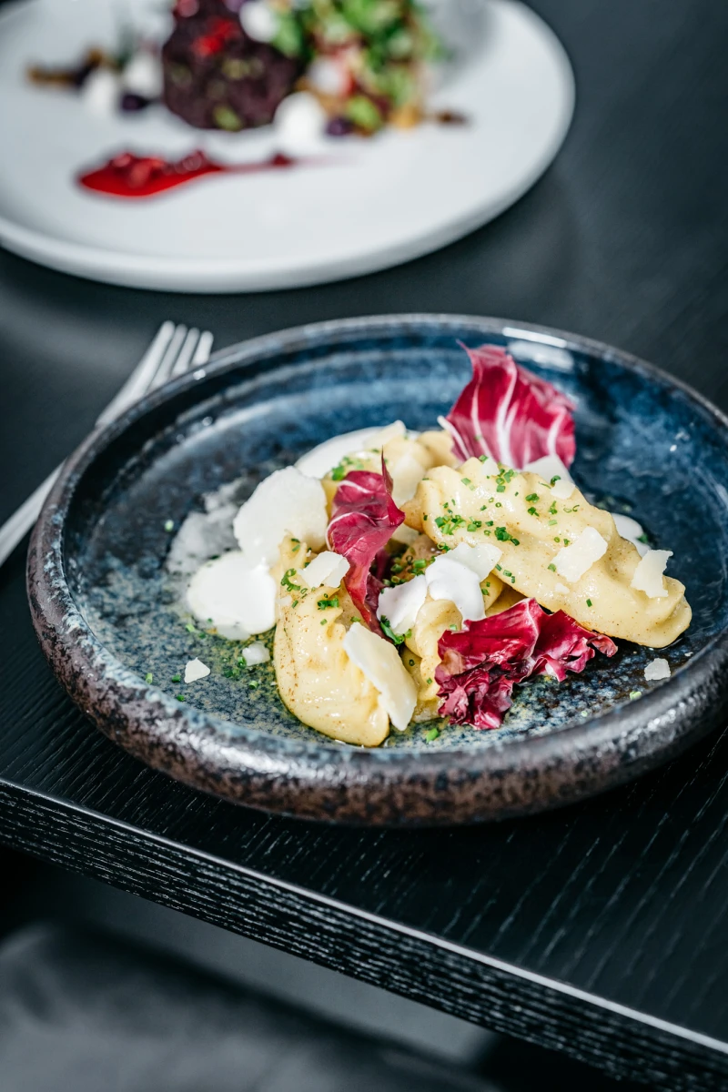 Feine Kartoffelgnocchi auf einem blauen Teller, garniert mit rosa Radicchio-Bl&auml;ttern und einer cremigen Sauce. Die Speise ist kunstvoll angerichtet und liegt auf einem dunklen, strukturierten Untergrund.