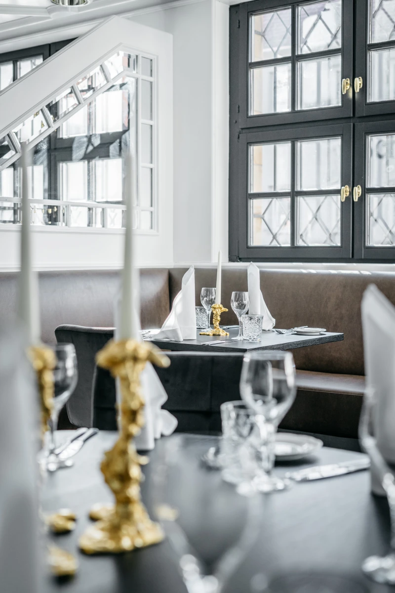 Gedeckter Tisch in elegantem Restaurant mit wei&szlig;en Tellern, Weingl&auml;sern und goldenen Kerzenhaltern. Gro&szlig;es Fenster mit Sprossenfenstern im Hintergrund, ged&auml;mpftes Licht und stilvolle Einrichtung.