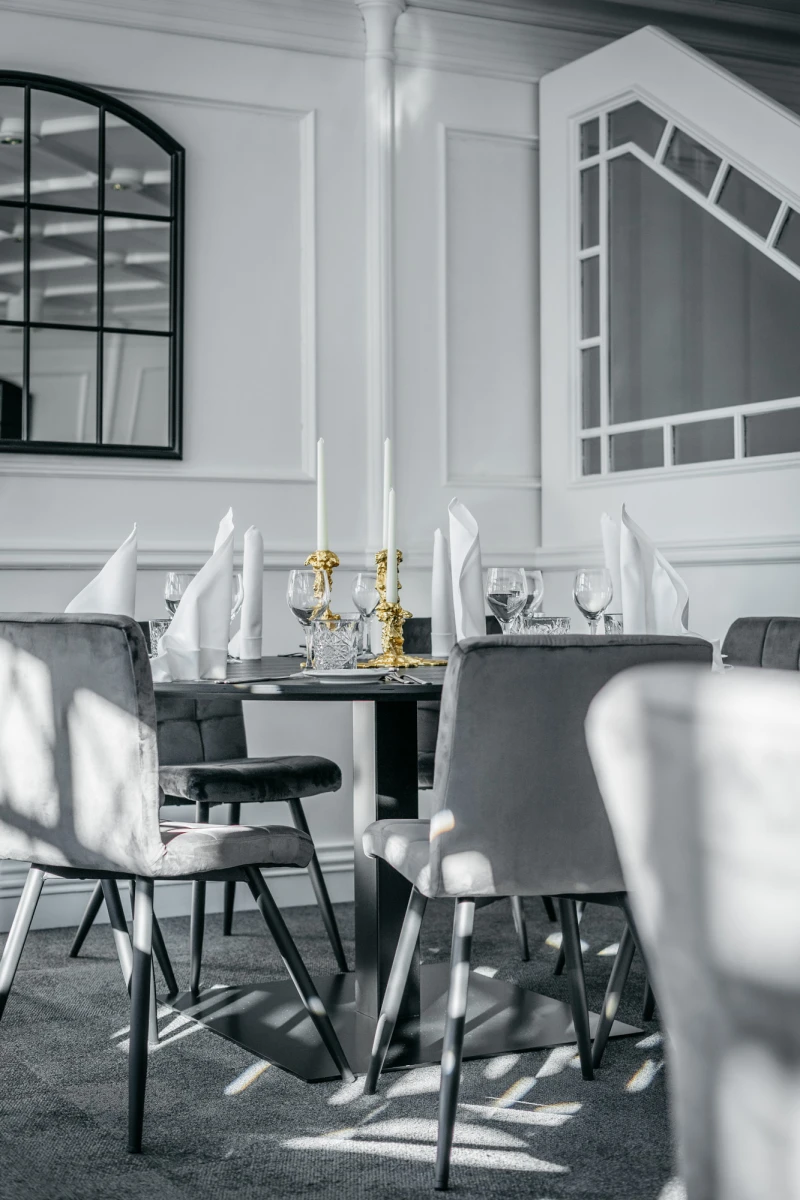 Eleganter Esszimmerbereich mit gedecktem Tisch, wei&szlig;en Stoffservietten und goldenen Kerzenhaltern. Moderne graue St&uuml;hle stehen um einen Tisch, umgeben von stilvollen wei&szlig;en W&auml;nden und gro&szlig;en Fenstern.