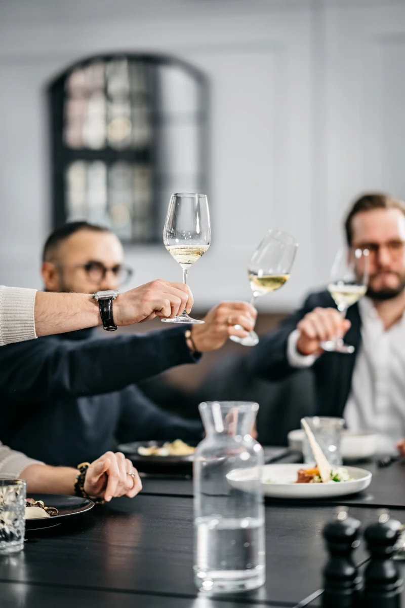 Drei Personen sto&szlig;en mit Weingl&auml;sern an einem Tisch an, im Hintergrund verschwommen eine gro&szlig;e Fensternische. Auf dem Tisch stehen Teller, Gl&auml;ser und eine Wasserkanne in einer eleganten, ged&auml;mpften Atmosph&auml;re.