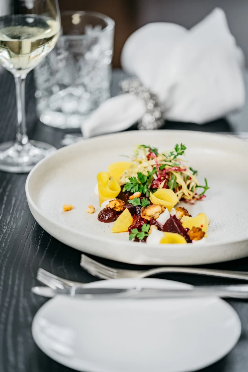 Elegantes Gericht auf wei&szlig;em Teller mit gelben und roten Elementen, garniert mit frischen Kr&auml;utern. Ein Glas Wei&szlig;wein und gestapelte wei&szlig;e Teller im Hintergrund deuten auf ein gehobenes Restaurant oder Dining-Erlebis hin.