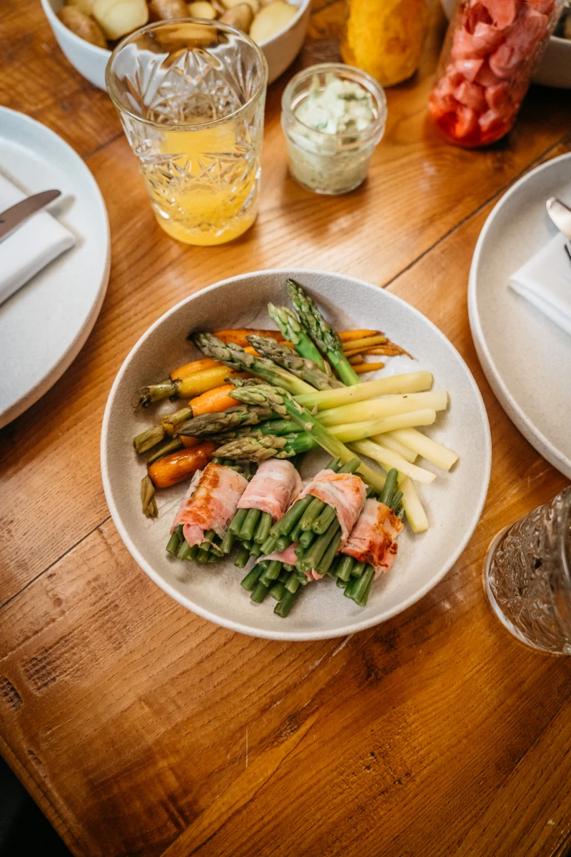 Ged&auml;mpfter Spargel, umwickelt von Schinken, serviert auf einem wei&szlig;en Teller, daneben gelbes Getr&auml;nk und Dekoration auf einem rustikalen Holztisch. Verschiedene Gem&uuml;sesorten wie gr&uuml;ner und gelber Spargel sowie Karotten sind appetitlich angerichtet.