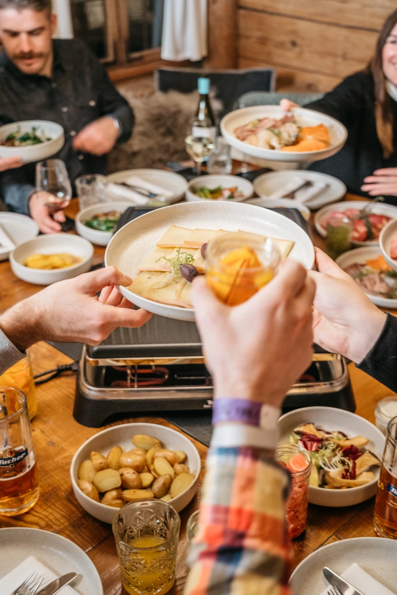 Gem&uuml;tliche Raclette-Mahlzeit mit verschiedenen Tellern, Beilagen und Zutaten auf einem Holztisch. Eine Hand h&auml;lt eine Pfanne mit geschmolzenem K&auml;se und einer Orangenscheibe, w&auml;hrend im Hintergrund weitere Personen am Tisch sitzen.