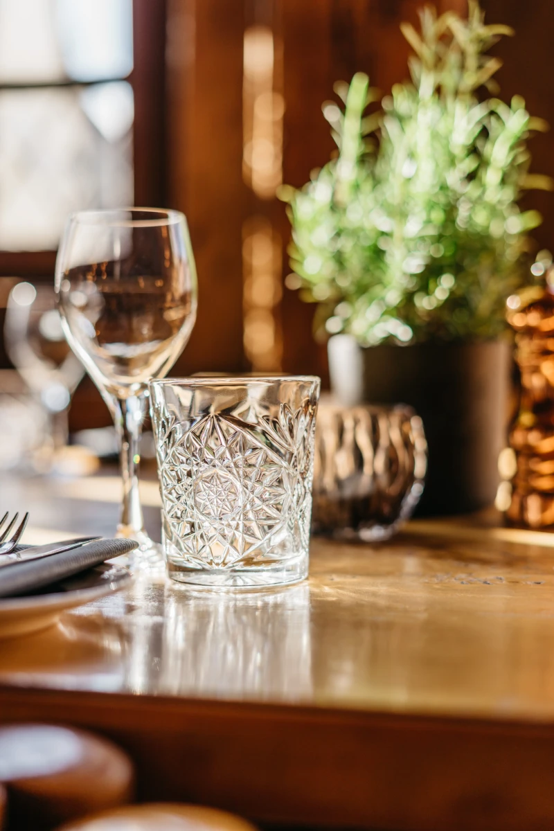 Geschliffenes Whiskyglas auf einem goldenen Holztisch mit unscharfem Hintergrund, im Hintergrund eine kleine gr&uuml;ne Pflanze in einem Topf.