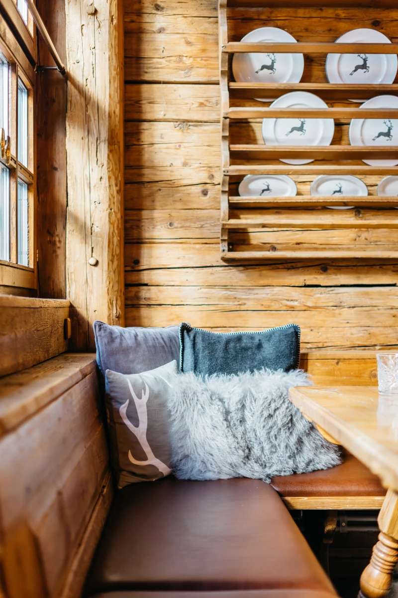 Gem&uuml;tliche Holzstube mit rustikaler Sitzbank, weichen Kissen in Blau- und Graut&ouml;nen und einem Tellerregal an der Holzwand. Warmes Sonnenlicht f&auml;llt durch ein Holzfenster und beleuchtet den einladenden, traditionellen Wohnbereich.