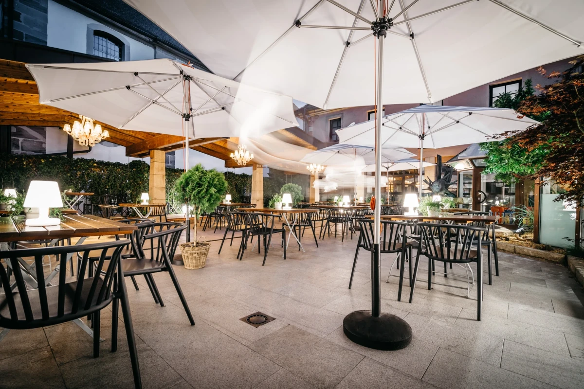 Heller Biergarten mit wei&szlig;en Sonnenschirmen und schwarzen Metallst&uuml;hlen, umgeben von Gr&uuml;npflanzen und dekorativen Leuchten. Der gepflasterte Au&szlig;enbereich bietet eine einladende Atmosph&auml;re f&uuml;r G&auml;ste zum Entspannen und Genie&szlig;en.