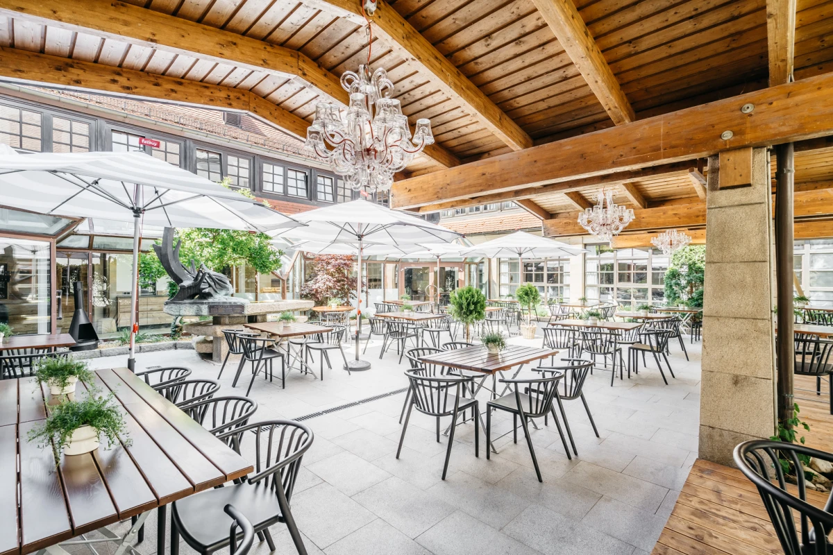 Ger&auml;umige Terrasse mit Holzdachstuhl, ausgestattet mit schwarzen Metalltischen und -st&uuml;hlen sowie wei&szlig;en Sonnenschirmen. Kristalll&uuml;ster h&auml;ngt von der Holzdecke, und die Einrichtung wirkt hell und einladend mit Blick auf einen Gartenbereich.