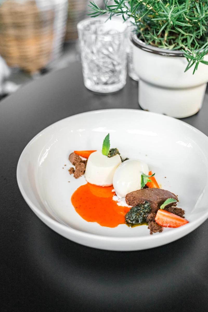 Raffiniertes Gourmet-Gericht auf wei&szlig;em Teller mit zart angerichtetem Fleisch, cremigem wei&szlig;em Klecks, orangefarbener So&szlig;e und gr&uuml;nen Kr&auml;utern, im Hintergrund ein Rosmarin-Topf und ein strukturiertes Glas.