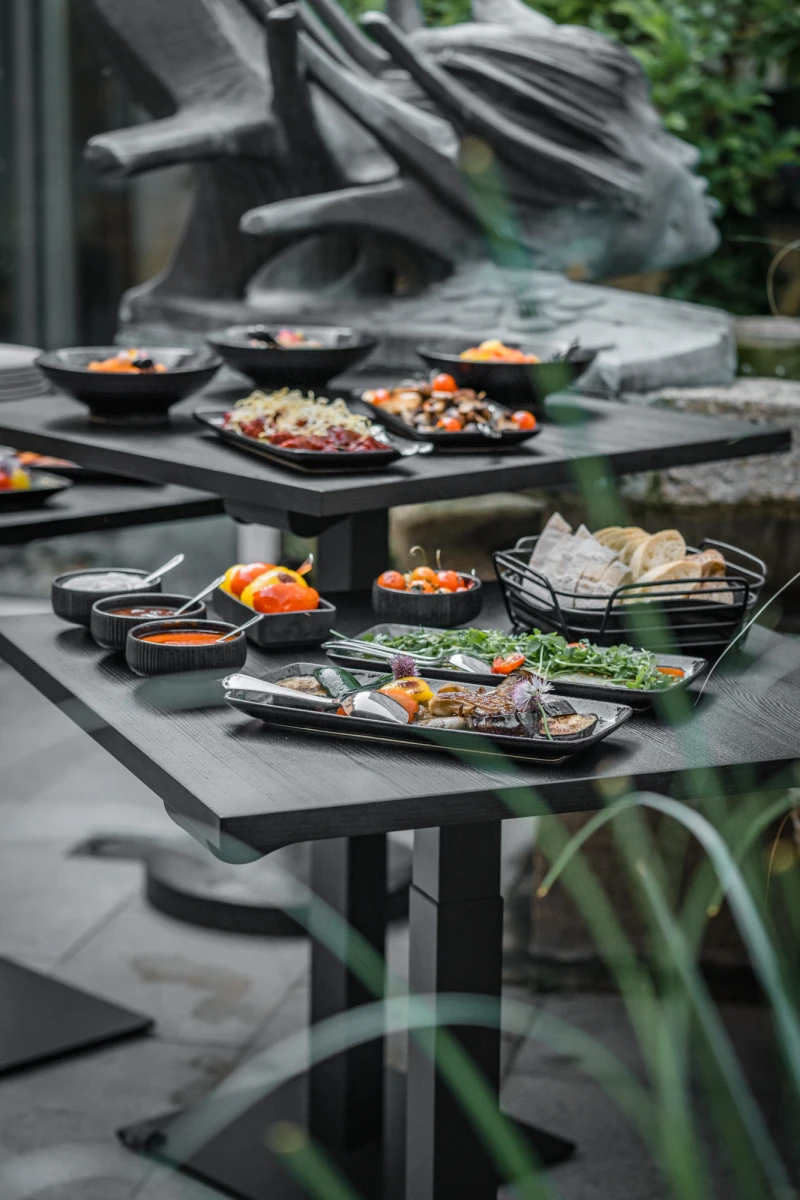 Gedeckter Tisch mit verschiedenen Gerichten in schwarzen Schalen und Tellern, angerichtet auf dunklen Tischen im Freien mit verschwommenen Gr&uuml;npflanzen im Vordergrund und einem steinernen Hintergrund.