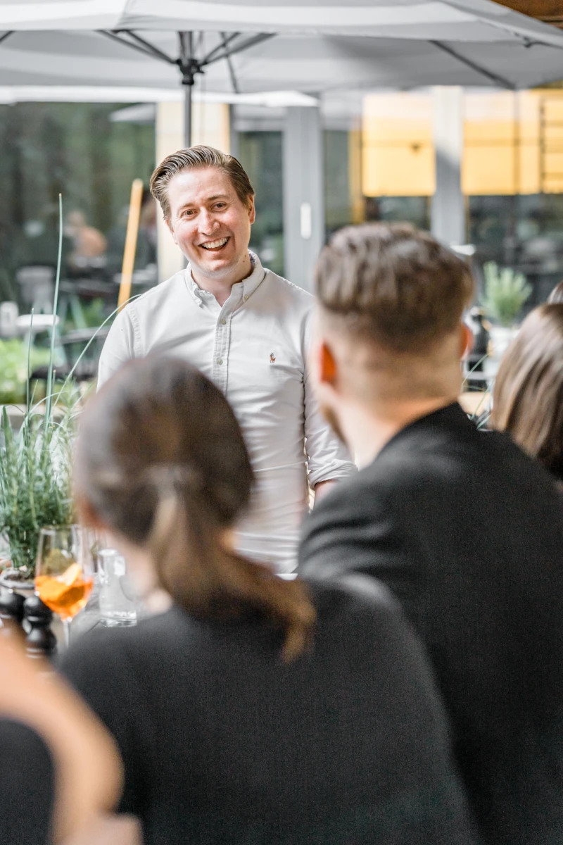 Ein l&auml;chelnder Mann in hellem Hemd steht auf einer Terrasse und unterh&auml;lt sich mit Kollegen oder G&auml;sten unter einem wei&szlig;en Sonnenschirm in einer gr&uuml;nen, modernen Umgebung.