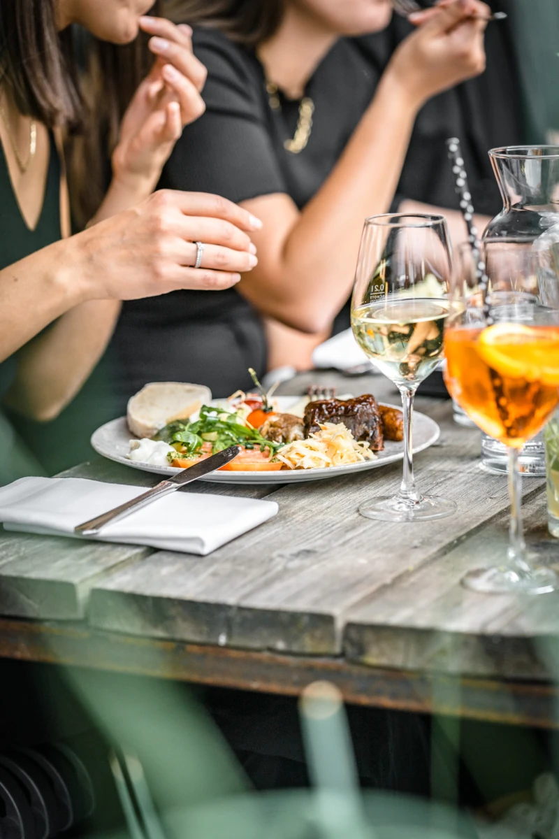 Gedeck auf einem rustikalen Holztisch mit einem Teller Speisen, einem Glas Wei&szlig;wein und einem orangefarbenen Cocktail. Personen in dunkler Kleidung essen gemeinsam, nur H&auml;nde und Ausschnitt sichtbar.