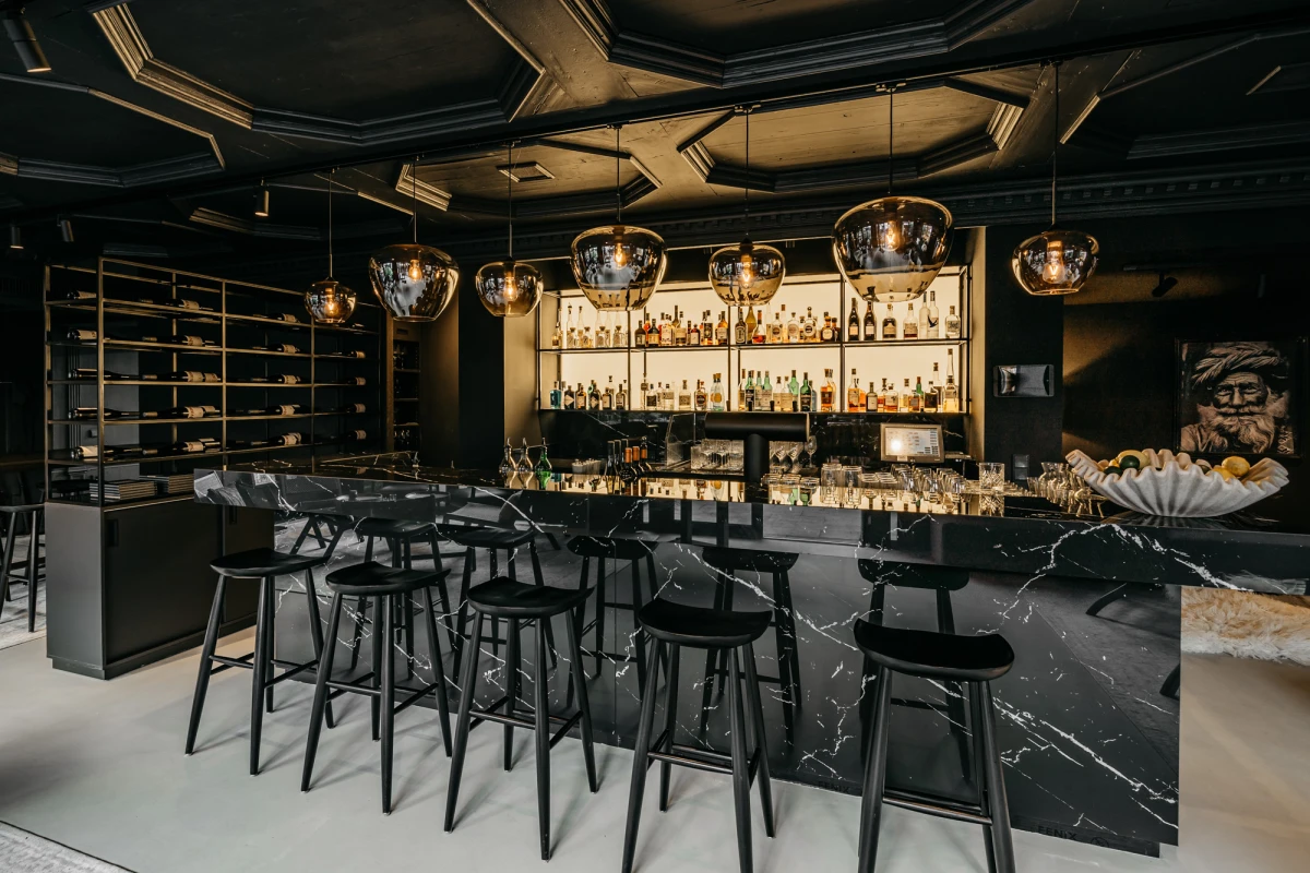 Stilvolle Bar mit elegantem, schwarzem Marmor-Tresen und hohen Barhockern, goldenen H&auml;ngelampen und einer beeindruckenden Spirituosensammlung an der R&uuml;ckwand. Die Decke ist aufwendig gestaltet und der Raum strahlt eine moderne, luxuri&ouml;se Atmosph&auml;re aus.