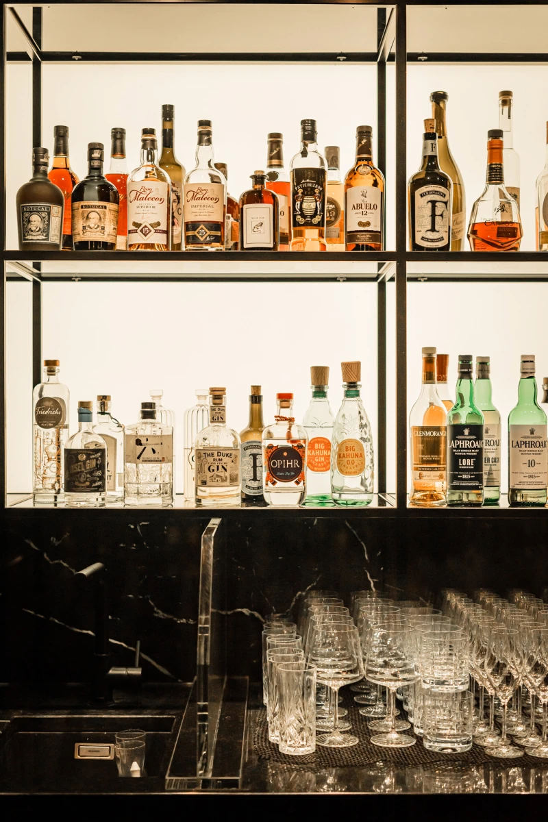 Elegante Hausbar mit zwei Regalbrettern voller verschiedener Spirituosen und Flaschen in warmen T&ouml;nen. Darunter eine Theke mit gestapelten Whiskygl&auml;sern und Kristallgl&auml;sern. Die Anordnung ist symmetrisch und ordentlich, mit einer Vielzahl von Alkoholen in unterschiedlichen Formen und Farben.