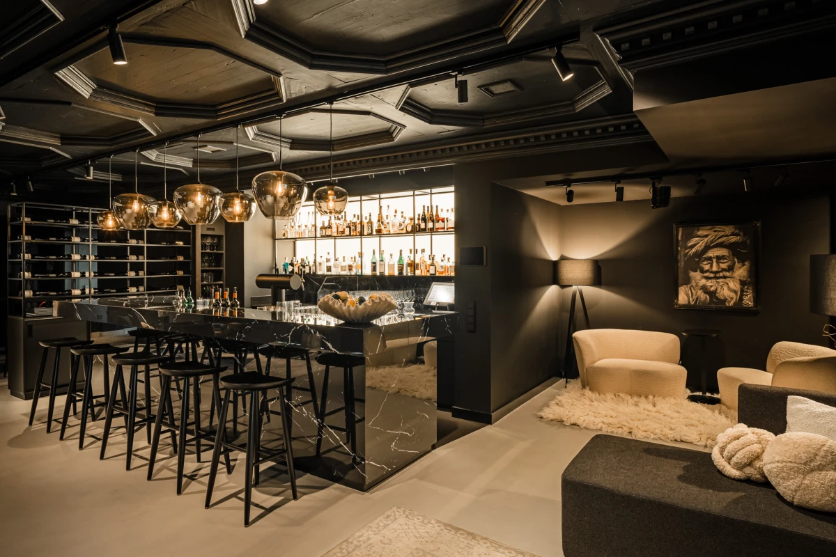 Stilvolle Bar mit elegantem schwarzem Tresen, goldenen H&auml;ngelampen und Barhocker. Rechts ein gem&uuml;tlicher Sitzbereich mit beigen Polsterm&ouml;beln und einem Kunstport&auml;t an der Wand. Die Atmosph&auml;re ist edel und modern mit ged&auml;mpftem Licht und hochwertigem Interieur.