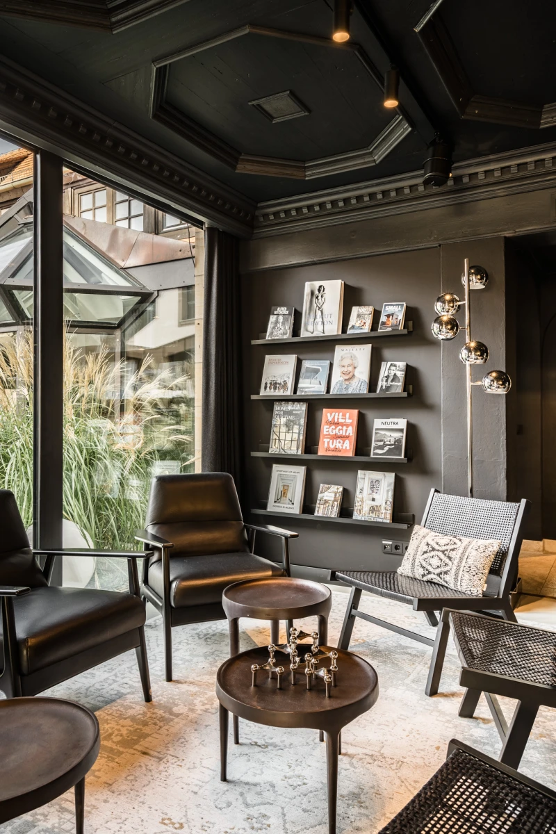 Stilvoller Lesebereich mit Panoramafenstern und schwarzen W&auml;nden, ausgestattet mit Ledersesseln, Couchtischen und einer Kunstwand mit sorgsam arrangierten B&uuml;chern und Bildern. Dezente Beleuchtung und ein modernes, minimalistisches Design schaffen eine einladende Atmosph&auml;re.