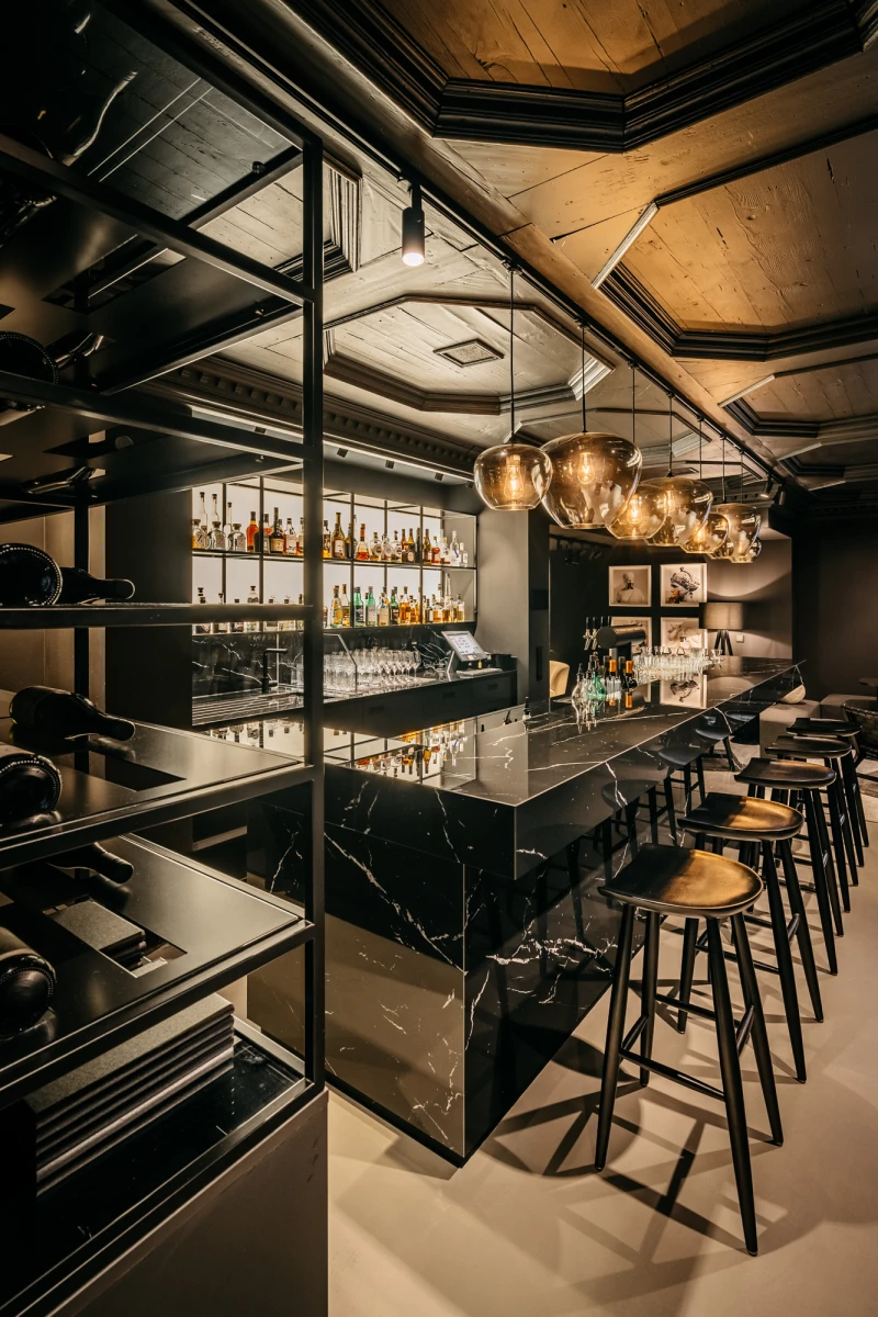 Elegante Bar mit schwarzer Marmorbartheke, goldenen H&auml;ngelampen und Barhockern. Dahinter offene Regale mit verschiedenen Spirituosen und Getr&auml;nken, umrahmt von schwarzen Metallgestellen. Die Decke ist in warmen Holzt&ouml;nen gehalten, was eine gediegene und moderne Atmosph&auml;re schafft.