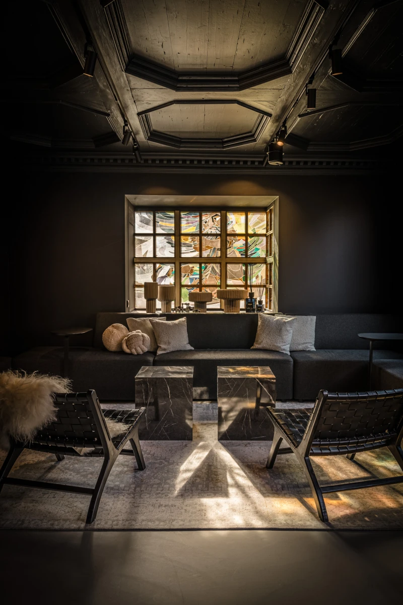 Rustikale Lounge mit Holzm&ouml;beln und gro&szlig;em Fenster, das Tageslicht hereinl&auml;sst. Dunkle Lederst&uuml;hle und Holztisch bilden einen gem&uuml;tlichen Sitzbereich vor dem Fenster mit Ausblick auf B&auml;ume.