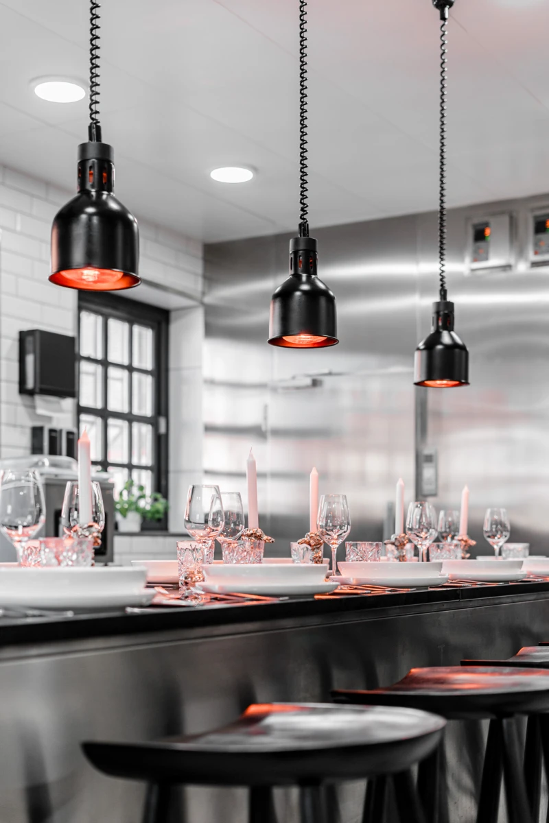 Moderne Bar-Theke mit schwarzen H&auml;ngelampen und rosa Weingl&auml;sern, dezent beleuchtet und elegant eingerichtet. Stahlgraue Oberfl&auml;chen und gro&szlig;e Fenster verleihen dem Raum eine zeitgen&ouml;ssische Atmosph&auml;re.