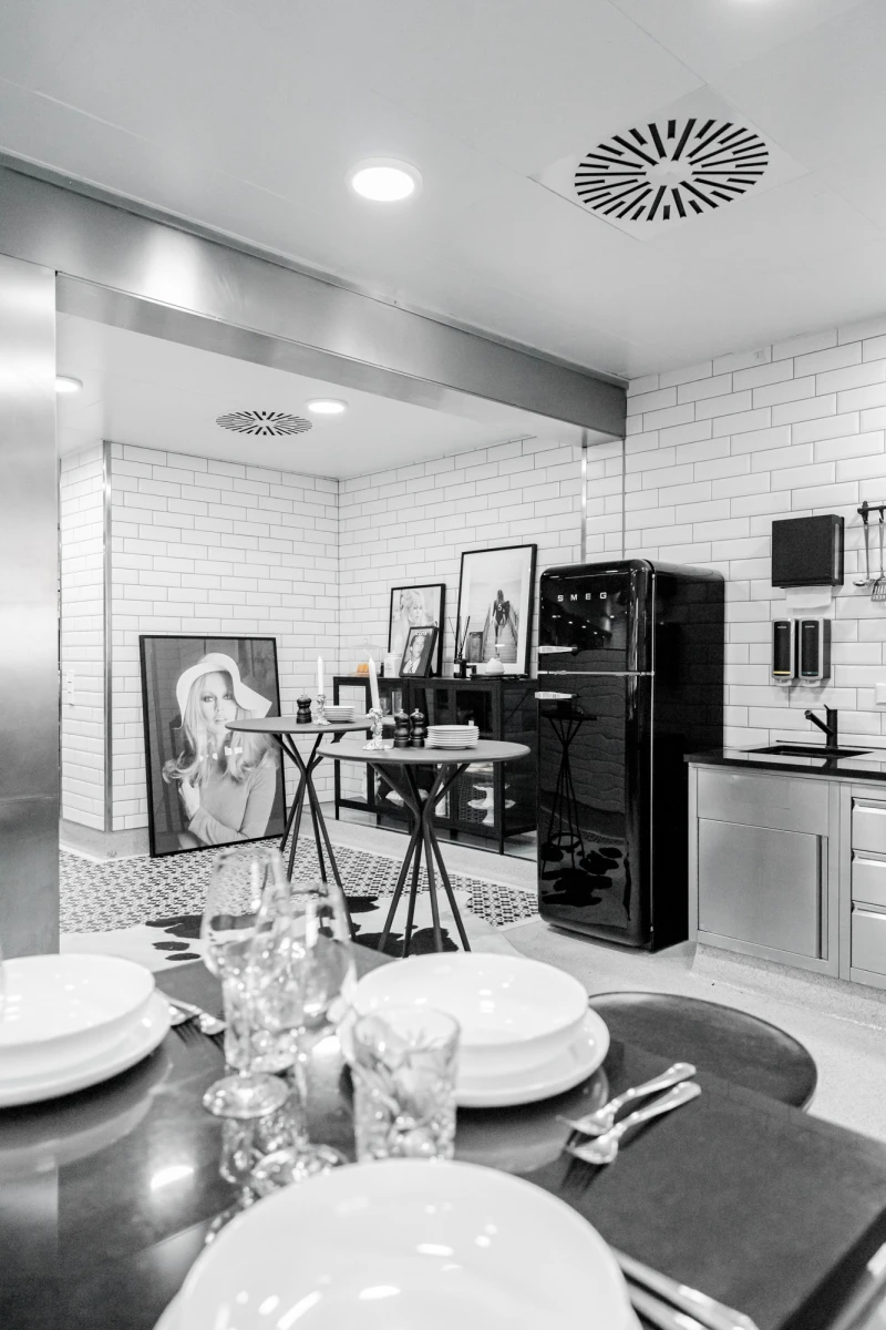 Moderne K&uuml;che mit schwarzer Retro-K&uuml;hlschrank, Ziegelfliesen und gedecktem Tisch im Vordergrund. Kunstvolle Atmosph&auml;re mit Gem&auml;lde und Rahmen, die an einer Wand lehnen. Dezentes Licht und klare Linien pr&auml;gen den minimalistischen Raum.
