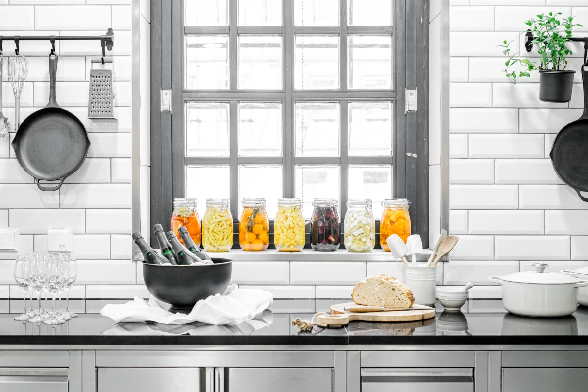 Moderne K&uuml;che mit wei&szlig;en Fliesen und grauen Akzenten, Einmachgl&auml;ser mit Obst oder Gew&uuml;rzen auf der Fensterbank, K&uuml;chenutensilien und Schneidebrett auf der Arbeitsplatte, rustikale und minimalistische Einrichtung.