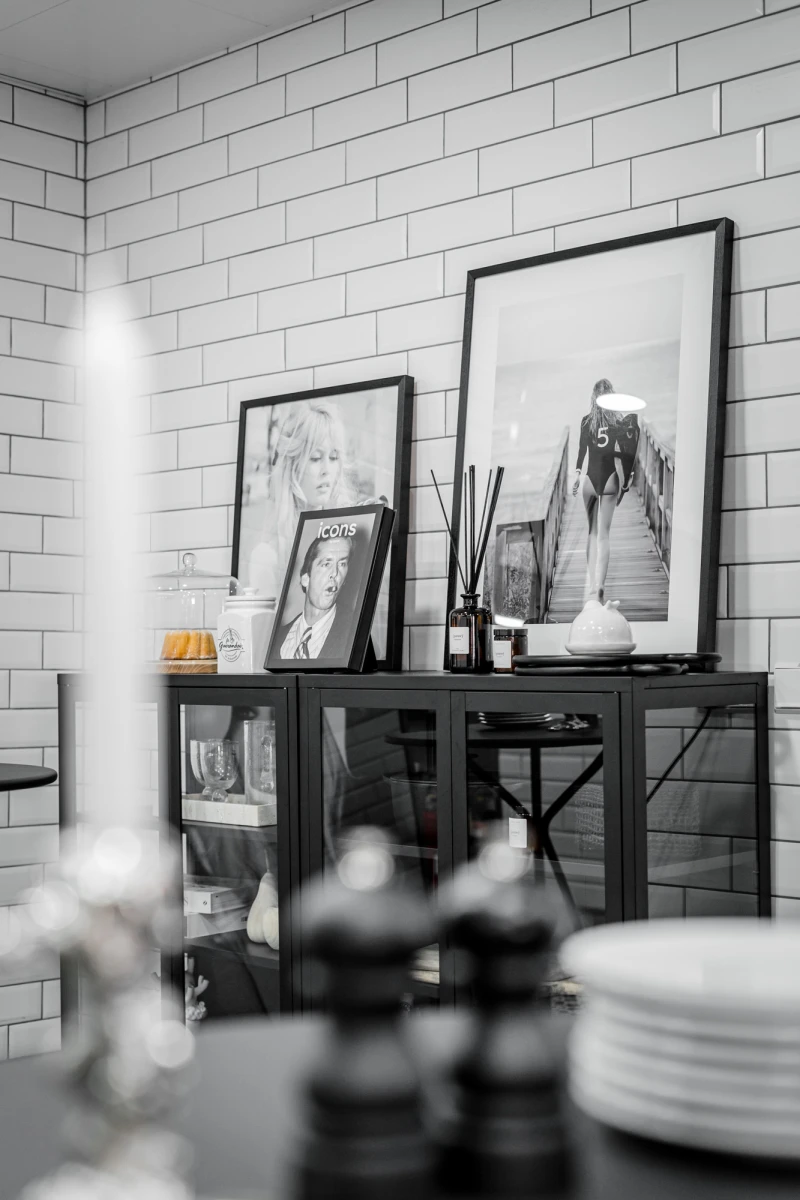 Schwarzwei&szlig;es Stillleben einer K&uuml;chen-Sideboard-Ecke mit gerahmten Kunstdrucken und Dekorationsobjekten vor einer wei&szlig;en Ziegelmauer. Auf dem Sideboard stehen verschiedene Bilderrahmen, Kerzen und Dekoration, im Vordergrund sind unscharf Geschirr und Gegenst&auml;nde zu sehen.
