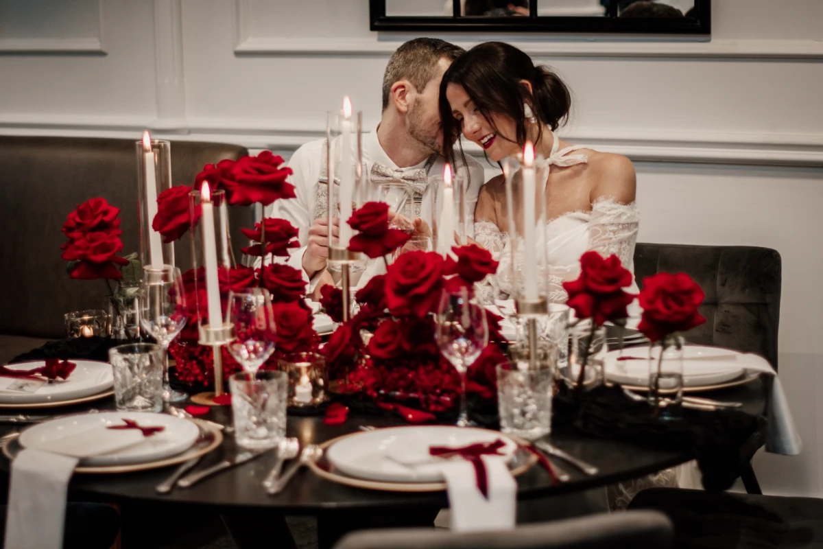Festlich gedeckter Tisch mit roten Rosen und brennenden Kerzen, zwei Personen in Hochzeitskleidung sitzen nah beieinander und teilen einen intmen Moment an einem eleganten Esstisch mit wei&szlig;em Geschirr und Kristallgl&auml;sern.