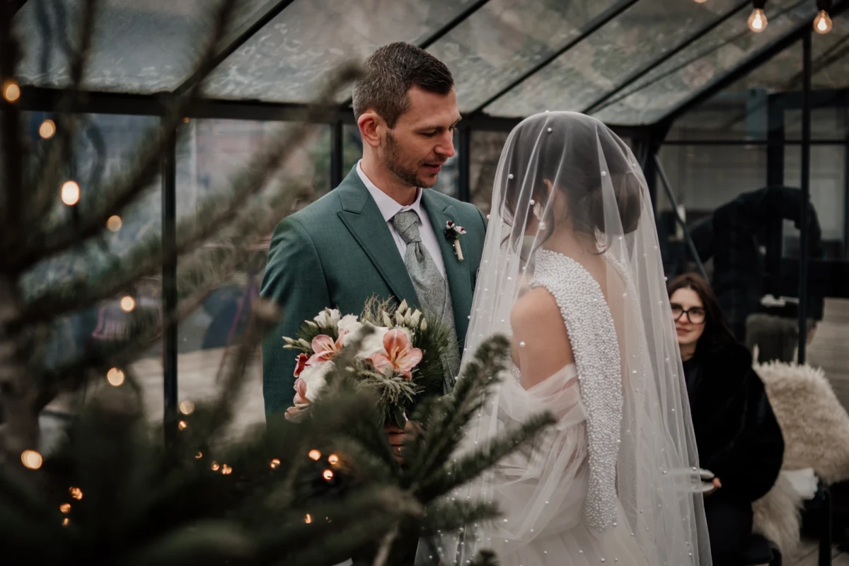 Brautpaar in einem Wintergarten mit Lichterketten, der Br&auml;utigam in einem gr&uuml;nen Anzug und die Braut in einem wei&szlig;en Kleid mit Schleier, w&auml;hrend sie einen Blumenstrauss h&auml;lt.