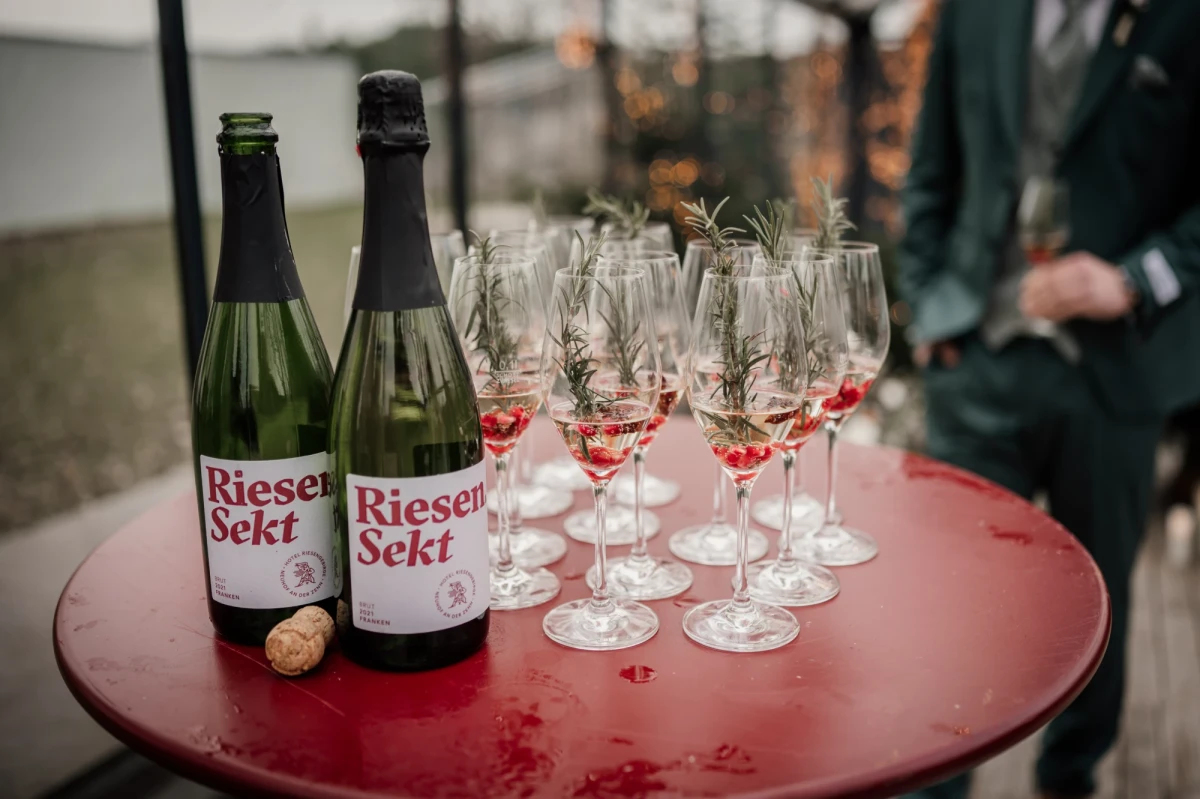 Zwei Flaschen Riesensekt auf einem roten Tisch mit mehreren Sektgl&auml;sern, die mit roten Details verziert sind. Im Hintergrund verschwommene Umrisse einer Person und einer Umgebung.