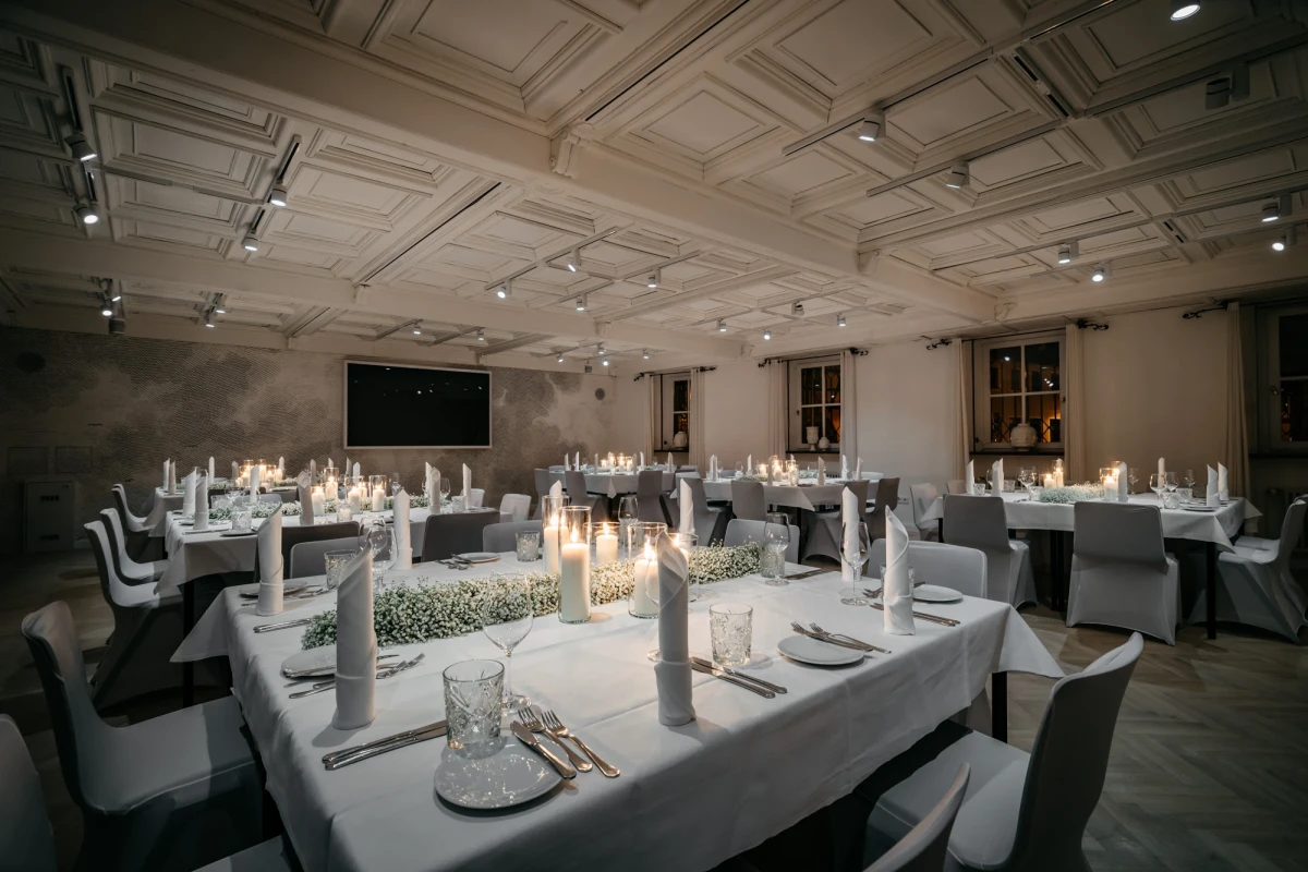 Festlich gedeckte Tische mit wei&szlig;en Tischdecken und brennenden Kerzen in einem eleganten Raum mit Kassettendecke und dezenter Beleuchtung. Graue St&uuml;hle und eine ged&auml;mpfte, intime Atmosph&auml;re pr&auml;gen den Raum, der f&uuml;r ein besonderes Dinner vorbereitet scheint.
