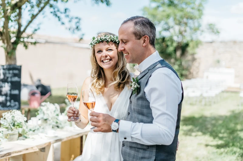 Braut mit Blumenkranz und Br&auml;utigam in Weste stehen an einem Hochzeitstag auf einer Wiese, halten Sektgl&auml;ser und l&auml;cheln gl&uuml;cklich miteinander.