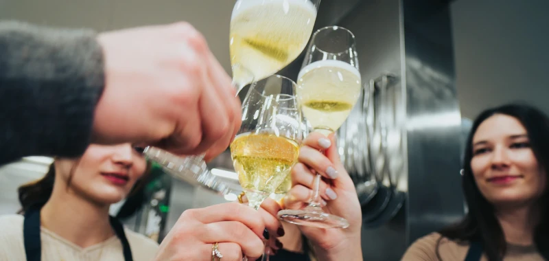 Mehrere H&auml;nde sto&szlig;en mit Champagnergl&auml;sern an. Die Gl&auml;ser sind gef&uuml;llt mit gelblichem Champagner und werden vor einem unscharfen Hintergrund gehoben.