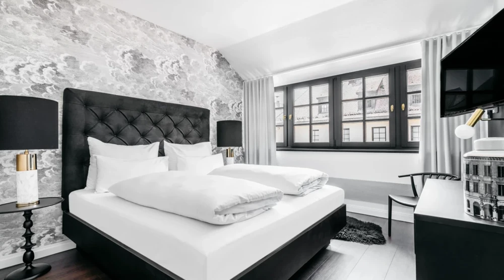 Ein elegant eingerichtetes Hotelzimmer mit einem schwarzen gepolsterten Bett, wei&szlig;er Bettw&auml;sche und einem gro&szlig;en Fenster. Graue Tapete und schwarze Akzente wie Nachttischlampen und Bodenbelag verleihen dem Raum einen modernen, kontrastierenden Look.