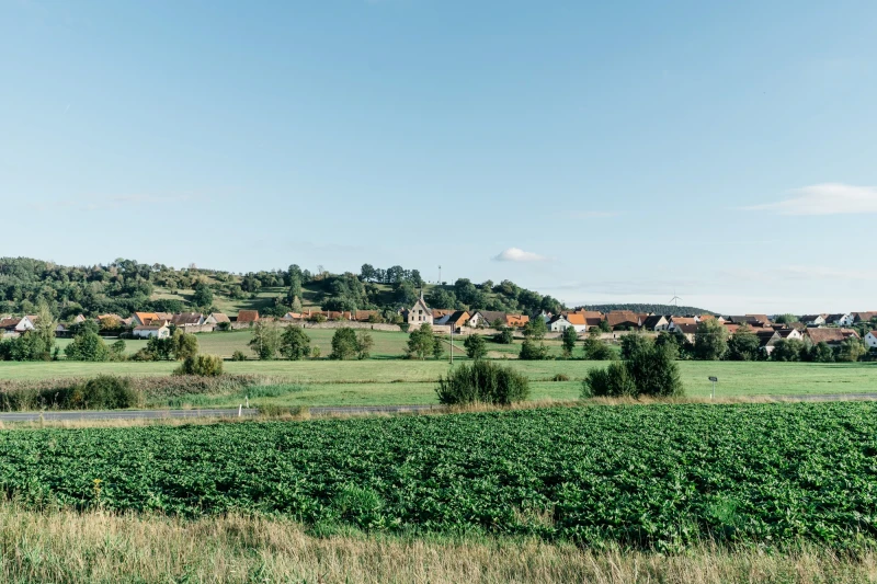 Gr&uuml;ne Landschaft mit Feldern im Vordergrund und einem Dorf mit roten D&auml;chern am Horizont. Sanfte H&uuml;gel und blauer Himmel bilden eine friedliche l&auml;ndliche Szene.