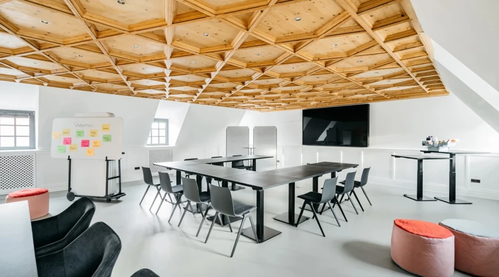 Moderner Besprechungsraum mit Holzbalkendecke, gro&szlig;em wei&szlig;em Tisch, schwarzen St&uuml;hlen und Fernsehbildschirm. Buinte Haftnotizen an der Wand, rote und schwarze Sitzhocker im Raum verteilt. Helle, offene Atmosph&auml;re mit Dachfenster und minimalistischem Design.