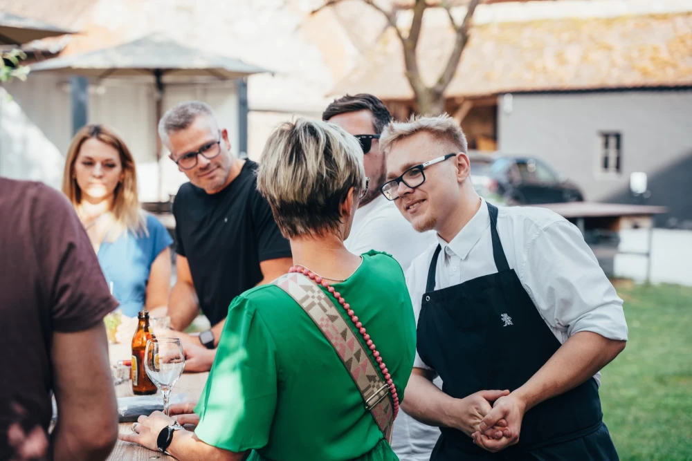 Gruppe von Menschen unterh&auml;lt sich auf einem Hoffest, ein junger Mann in Sch&uuml;rze steht im Vordergrund und l&auml;chelt, w&auml;hrend andere Personen im Hintergrund Bier trinken und plaudern.