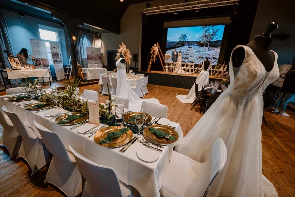 Hochzeitstafel mit wei&szlig; gedeckten Tischen, goldenen Tellern und Gr&uuml;ndekor, einem Brautkleid auf einer Schaufensterpuppe im Vordergrund und einer Pr&auml;sentation auf einer Leinwand im Hintergrund, die eine Hochzeitsveranstaltung in einem Veranstaltungsraum zeigt.