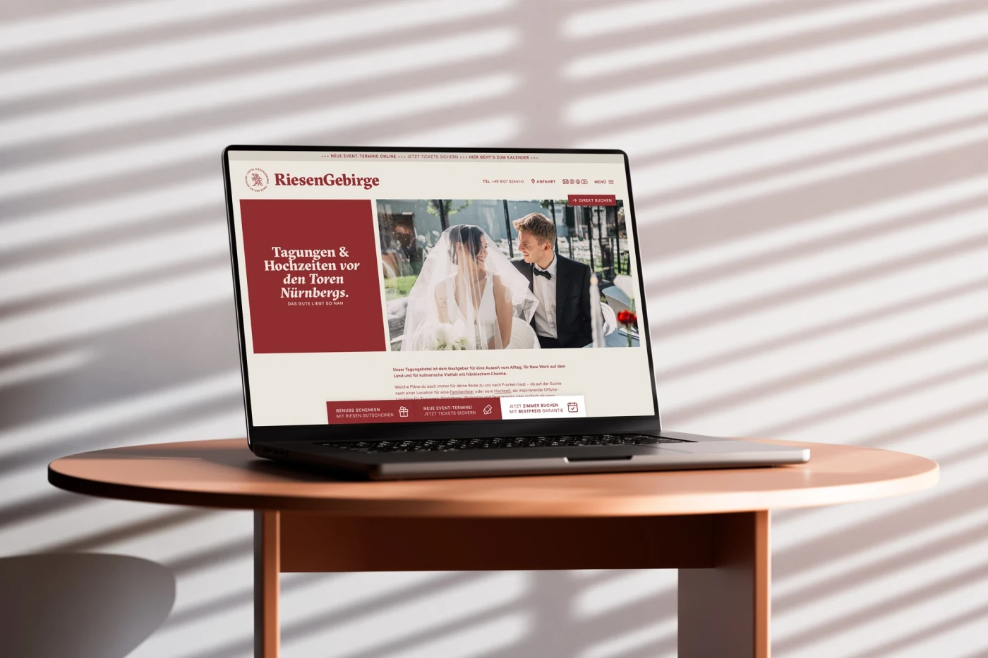 Laptop auf einem runden Holztisch, der die Website von Hotel Riesengebirge pr&auml;sentiert, vor einer Wand mit Lichtschatten-Streifen.