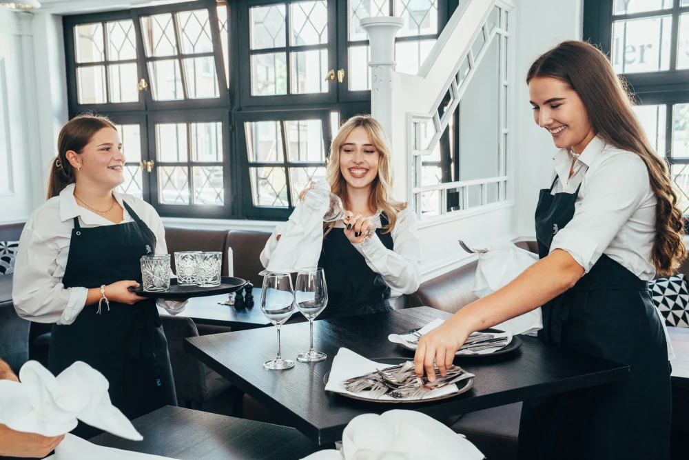 Drei junge Kellnerinnen in schwarzer Kochsch&uuml;rze und wei&szlig;er Bluse arbeiten gemeinsam in einer hellen, modernen K&uuml;che. Sie l&auml;cheln und bereiten Teller und Gl&auml;ser f&uuml;r den Service vor. Die Frauen strahlen Teamgeist und Freude an ihrer Arbeit aus.
