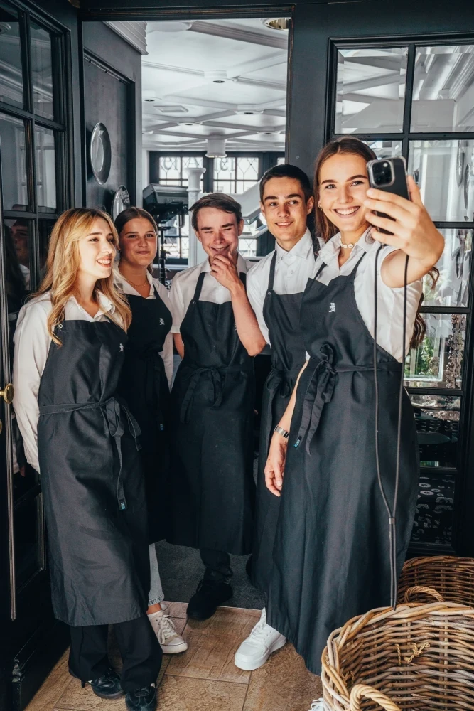 F&uuml;nf junge Mitarbeiter in grauen Sch&uuml;rzen stehen in einem modernen Caf&eacute; oder Restaurant und eine Person macht ein Selfie. Sie l&auml;cheln und posieren gemeinsam f&uuml;r ein Gruppenfoto, das die Teamstimmung und Arbeitsatmosph&auml;re einf&auml;ngt.